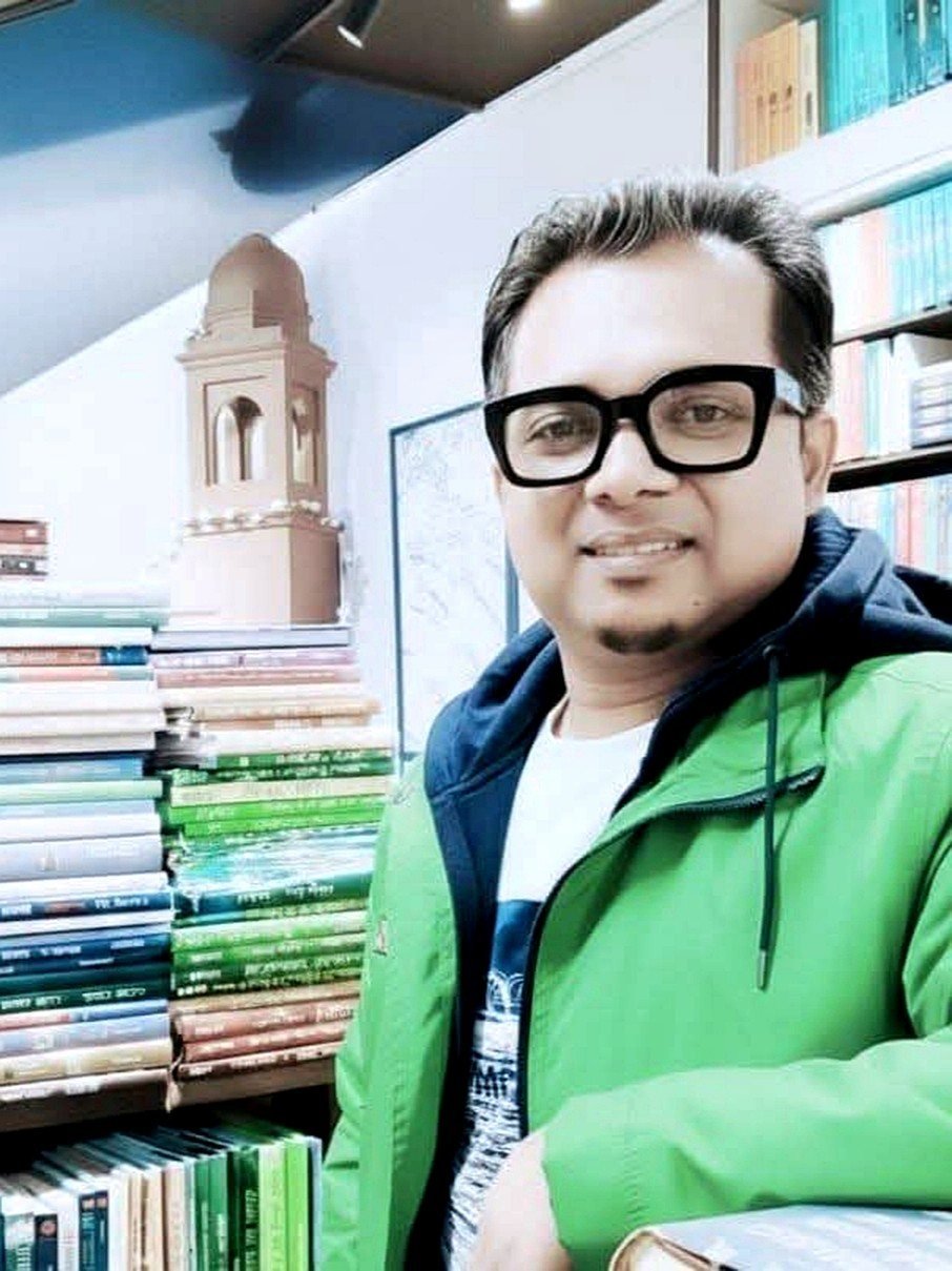 Masud Pathik, PhD, in a green jacket and black-framed glasses, among shelves and stacks of books